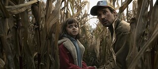 Stillframe from the Netflix Germany Series "The Signal" showing Florian David Fitz and Yuna Bennett in a corn field with a startled facial expression.