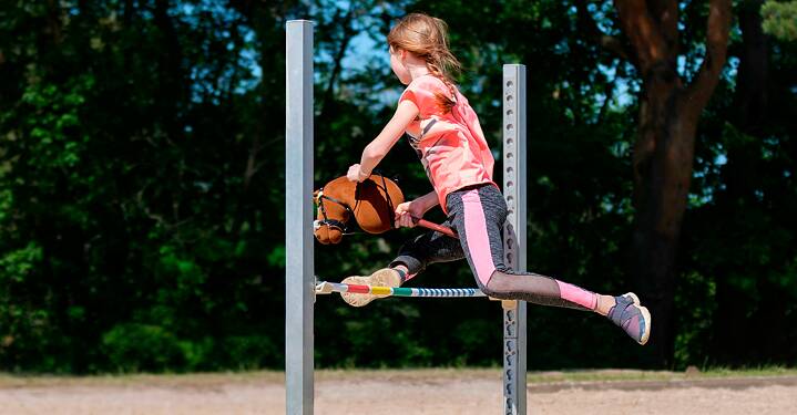 The picture shows a girl jumping over an obstacle with a hobbyhorse.