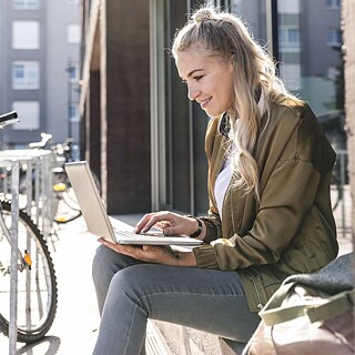 An einem sonnigen Tag sitzt eine junge Frau mit blonden langen Haaren auf einer Fensterbank und tippt auf ihrem aufgeklappten Laptop. Sie lächelt dabei. 