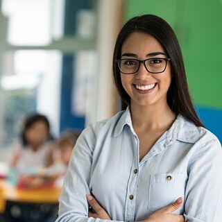 Porträt einer lächelnden jungen Lehrerin im Klassenzimmer. Im Hintergrund sitzen Schüler am Tisch. 