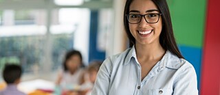 Porträt einer lächelnden jungen Lehrerin im Klassenzimmer. Im Hintergrund sitzen Schüler am Tisch. 