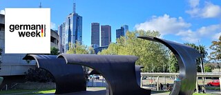 View of the Melbourne skyline on a sunny day. In the front there is a large black wavy sculpture. On the top left is the logo of German Week Melbourne