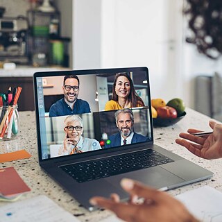 Videokonferenz im Homeoffice. Die Sprecherin gestikuliert; am Bildschirm sind weitere Konferenzteilnehmer zu sehen, sie lächeln freundlich und hören aufmerksam zu. Im Bildfokus sind die Gesprächspartner auf dem geteilten Monitor.