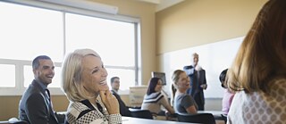 Ein Raum mit mehreren Personen in Geschäftskleidung, sie sitzen an unterschiedlichen Tischen, eine Dame im Vordergrund und ein unscharfer Hintergrund.