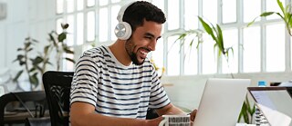 A young man with headphones sits at a desk and works on a laptop.