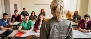 Rückansicht auf eine Lehrerin, die zu Schülern der Sekundarstufe im Klassenraum spricht.