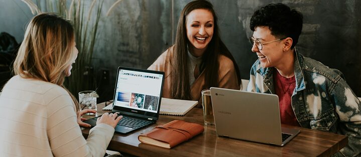 Three people sit at a table with laptops, laughing together. They appear relaxed, having a cheerful conversation in a cozy environment.