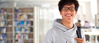 Ein Student mit grauem Kapuzenpullover und Brille steht lächelnd in der Bibliothek.