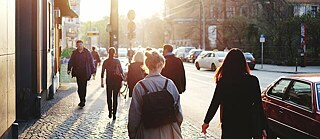 Fußgänger bewegen sich entlang einer Stadtstraße, umgeben von urbanen Gebäuden und Geschäften.