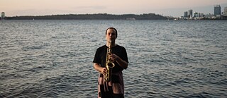 Sonic diary | Josten Myburgh - Josten spielt Saxophon, blickt direkt in die Kamera und steht knietief in einem Fluss. Es ist Abenddämmerung und der Himmel ist blau und rosa, mit ein paar kleinen Wolken. Im Hintergrund sind die Skyline von Boorloo und die grünen Hügel von Kaartagarup (King's Park) zu sehen. Im Vordergrund der Aufnahme befindet sich ein Mikrofon, das Josten aufnimmt.