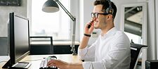 Ein Mann mit Brille sitzt an einem Schreibtisch, arbeitet am Computer und hat ein Headset am Kopf.