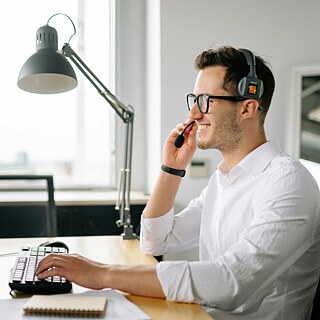 Ein Mann mit Brille sitzt an einem Schreibtisch, arbeitet am Computer und hat ein Headset am Kopf.