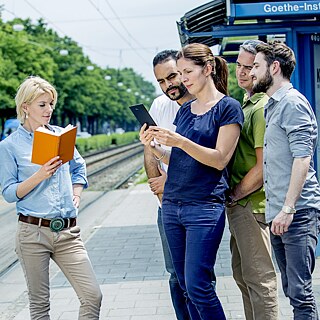 Fünf Personen stehen an einer Straßenbahnhaltestelle. Eine Frau hält ein Buch, während die anderen vier auf ein Tablet schauen. Im Hintergrund sind Gleise und Bäume zu sehen.