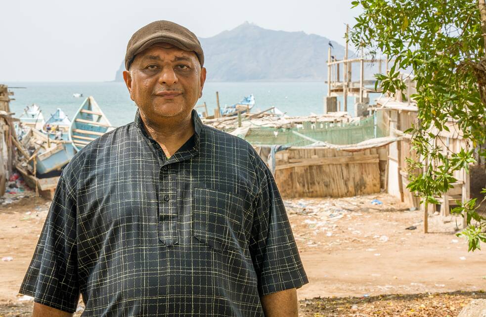 Kareem Hassan olha para a câmera. Ao fundo, pequenas botas de pescador antigas na costa do mar.