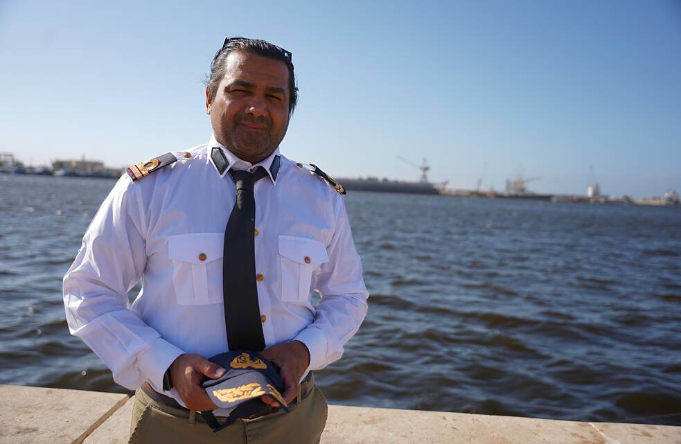 Wissam Hassan Al-Kawafi olha para a câmera. Atrás dele, você pode ver o oceano e a silhueta do Porto de Bengasi.i.