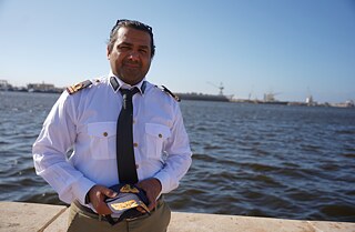 Wissam Hassan Al-Kawafi looks into the camera. Behind him you can see the ocean and the silhouette of the Port of Benghazi.