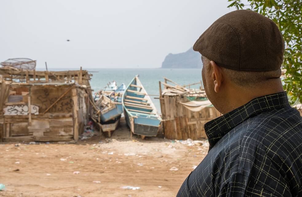 Kareem Hassan Mongy é visto de costas. Ele olha na direção do mar. Ao fundo, pequenas botas de pesca antigas na costa do mar.