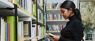 Young woman with long black hair stands in front of a bookshelf and leafs through a book.