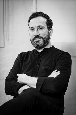 Black and white portrait of Eyal crossing his arms and smiling. Eyal is wearing a black top and glasses.  © Forensic Architecture 2020 Eyal Weizman B&W