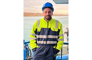 Bishoy Kamel Talaat looks into the camera. He weres his workers uniform in bright yellow and a blue security hat. Behind him you can see the ocean and the silhouette of the Canal.