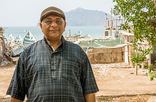 Kareem Hassan looks into the camera. You can see old small fishing boots on the shore of the sea in the background. 