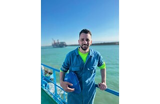 Bishoy Kamel Talaat olha para a câmera. Ele veste um uniforme azul. Atrás dele, é possível ver o oceano e a silhueta de uma embarcação no Canal.