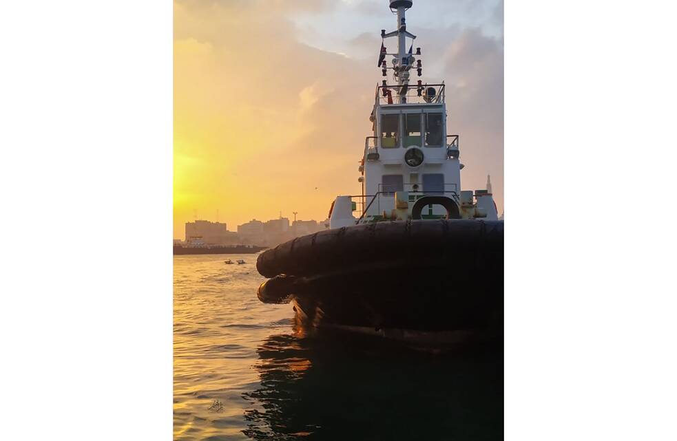 Parte de uma balsa no mar durante o pôr do sol. Ao fundo, a silhueta de uma cidade.