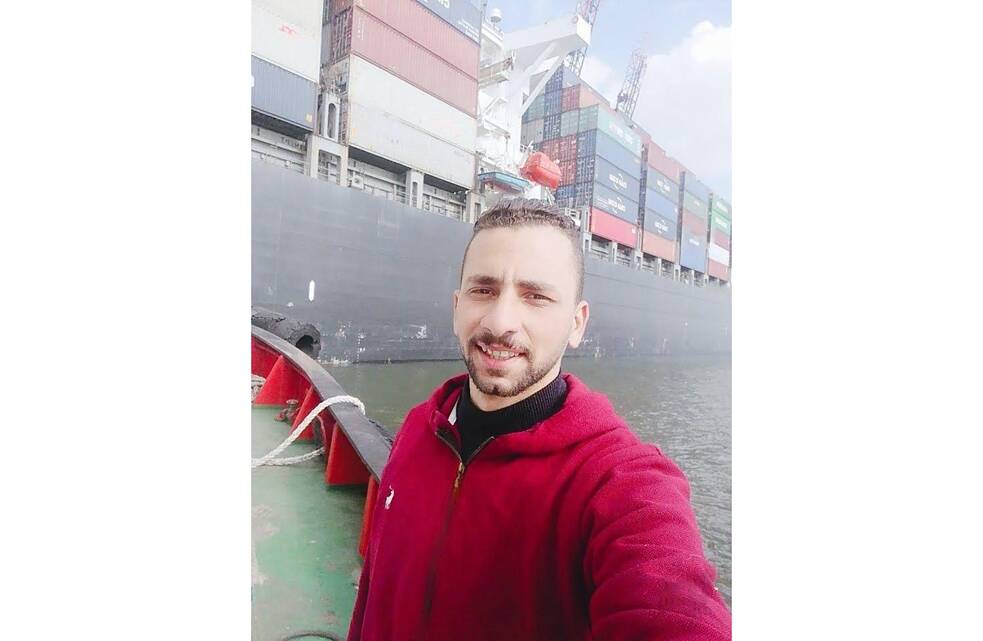 Bishoy Kamel Talaat olha para a câmera. Atrás dele, é possível ver um grande navio que transporta muitos contêineres grandes.