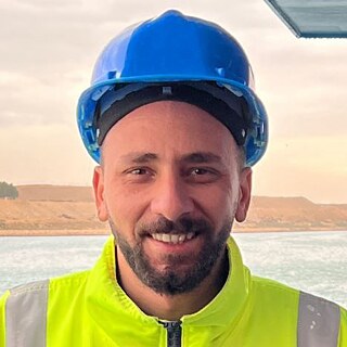 Bishoy Kamel Talaat looks into the camera. He weres his workers uniform in bright yellow and a blue security hat. Behind him you can see the ocean and the silhouette of the Canal.