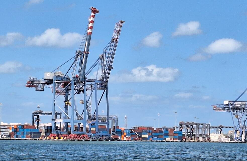 Grandes guindastes para carregar navios à direita, ao lado de vários contêineres de cores diferentes vistos do mar.