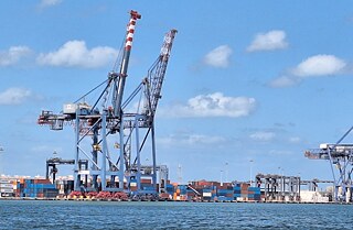 Große Kräne zum Beladen von Schiffen, rechts daneben viele verschiedenfarbige Container vom Meer aus gesehen.