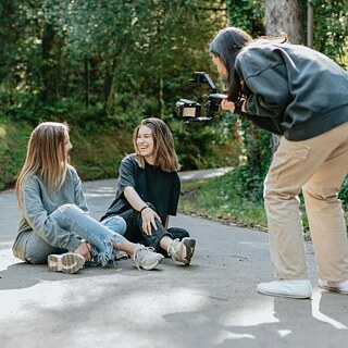 Two young women are seated on the ground, smiling as a young woman captures their joyful moment on camera.