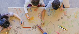 Eine Gruppe von Kindern sitzt an einem Tisch mit Papiertischdecke. Sie zeichnen mit Buntstiften umweltfreundliche Themen auf die Tischdecke. Aufnahme von oben.