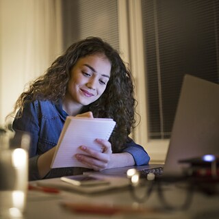 junge schwarzhaarige Studentin studiert zu Hause mit Laptop und macht sich Notizen im Notizbuch.