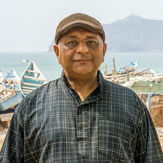 Kareem Hassan olha para a câmera. Ao fundo, pequenas botas de pesca antigas podem ser vistas na praia.