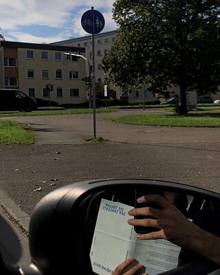 The picture shows a street with houses, traffic signs and trees photographed from a car. In the side mirror of the car, hands can be seen holding a telephone and the book Die Trauer der Tangente.