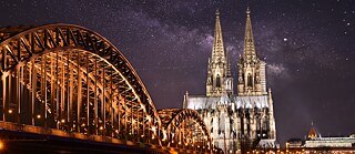 A stunning night view of the Cologne Cathedral and the Hohenzollern Bridge, illuminated against a starry sky, showcasing the iconic architecture and serene atmosphere.