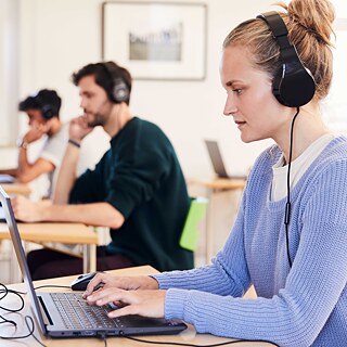 Drei Prüfungsteilnehmende mit Kopfhörern sitzen an Einzeltischen und arbeiten konzentriert an Laptops. 