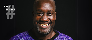 The author Musa Okwonga stands smiling against a jet-black background, wearing a purple glittering jumper. At the top left, the Vorzeichen logo, on the right, the book cover of In the End, it Was All About Love.