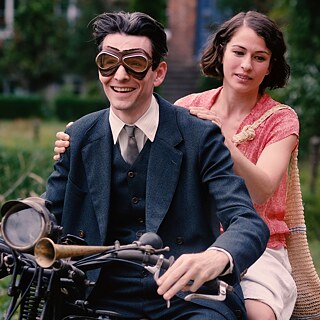 A man and a woman sit behind each other on a motorcycle in a rural setting