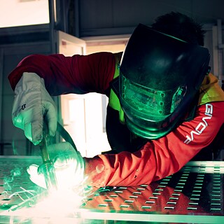 Person mit Schweißhelm und Handschuhen, konzentriert beim Schweißen einer Metallfläche.