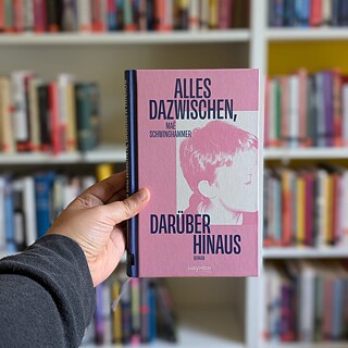 A hand holds the novel ‘Alles dazwischen, darüber hinaus’ in front of a bookshelf.