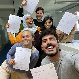 Selfie von Fero mit Tatjana, Sena, Noah und Amita. Sie stehen im Eingangsbereich des Goethe-Instituts und halten glücklich ihre Goethe-Zertifikate hoch.