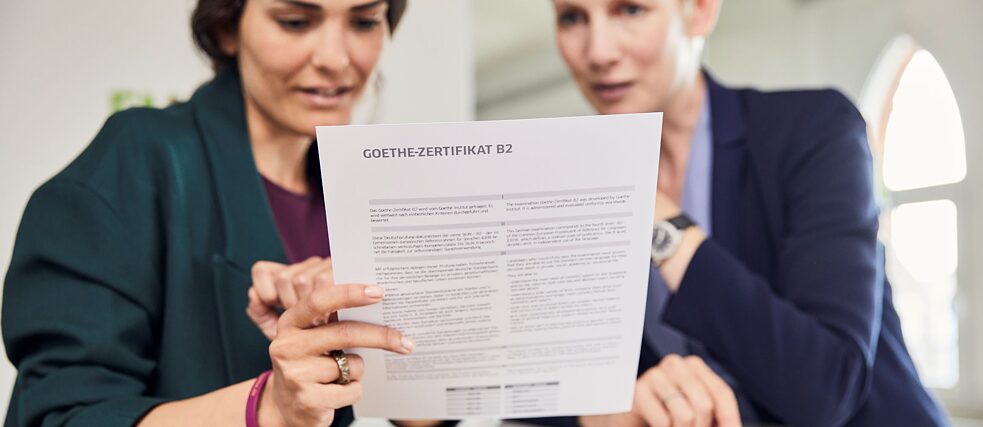 Gabriela hat die Prüfung bestanden. Sie hält das Goethe-Zertifikat B2 in der Hand und sichtet mit der Lehrkraft die Ergebnisse.