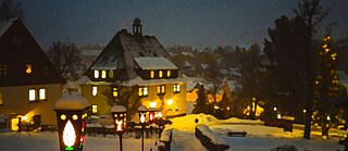 Ein mit Schnee bedecktes Dorf bei Nacht. Die Fenster leuchten gelb. Alles ist in warmes Licht getaucht. 