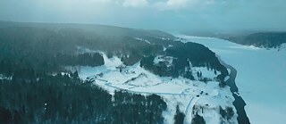 Wald, ein breiter Fluss und einige kleine Häuschen von oben im Winter. Alles ist mit Schnee bedeckt. Schneeflocken sind in der Luft. 