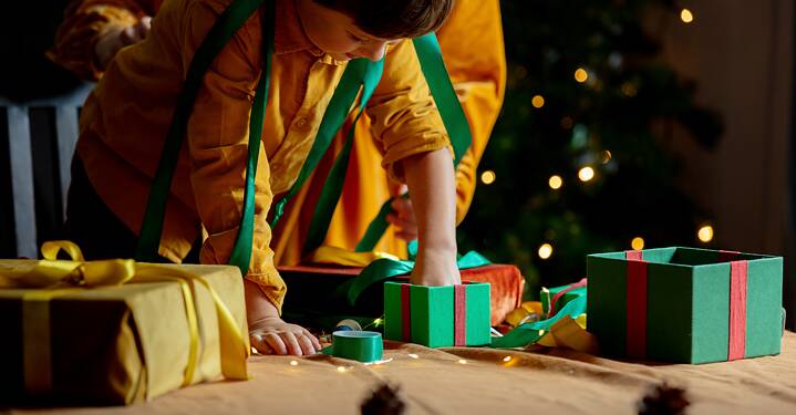 In the picture, a child is unwrapping a present from a box. There is a Christmas tree in the background. 