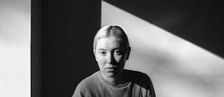 Black and white landscape image of Ellen wearing a knitted jumper and with her hair slicked back. A triangular shadow is on the wall behind her. 