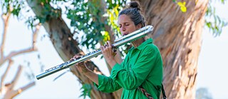 Sonic diary | Sabine Vogel - Sabine spielt Bassflöte und trägt dabei ein grünes Kleid und eine gewebte Tasche, die viele Farben hat: rosa, blau, gold, weiß und schwarz. Sie steht auf einer Terrasse, und hinter ihr steht ein Baum mit einem sehr großen Stamm und leuchtend grünen Blättern sowie ein Blick in die Ferne.