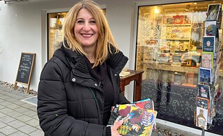 A blonde woman holds three books. She smiles. In the background there is a bookstore. 